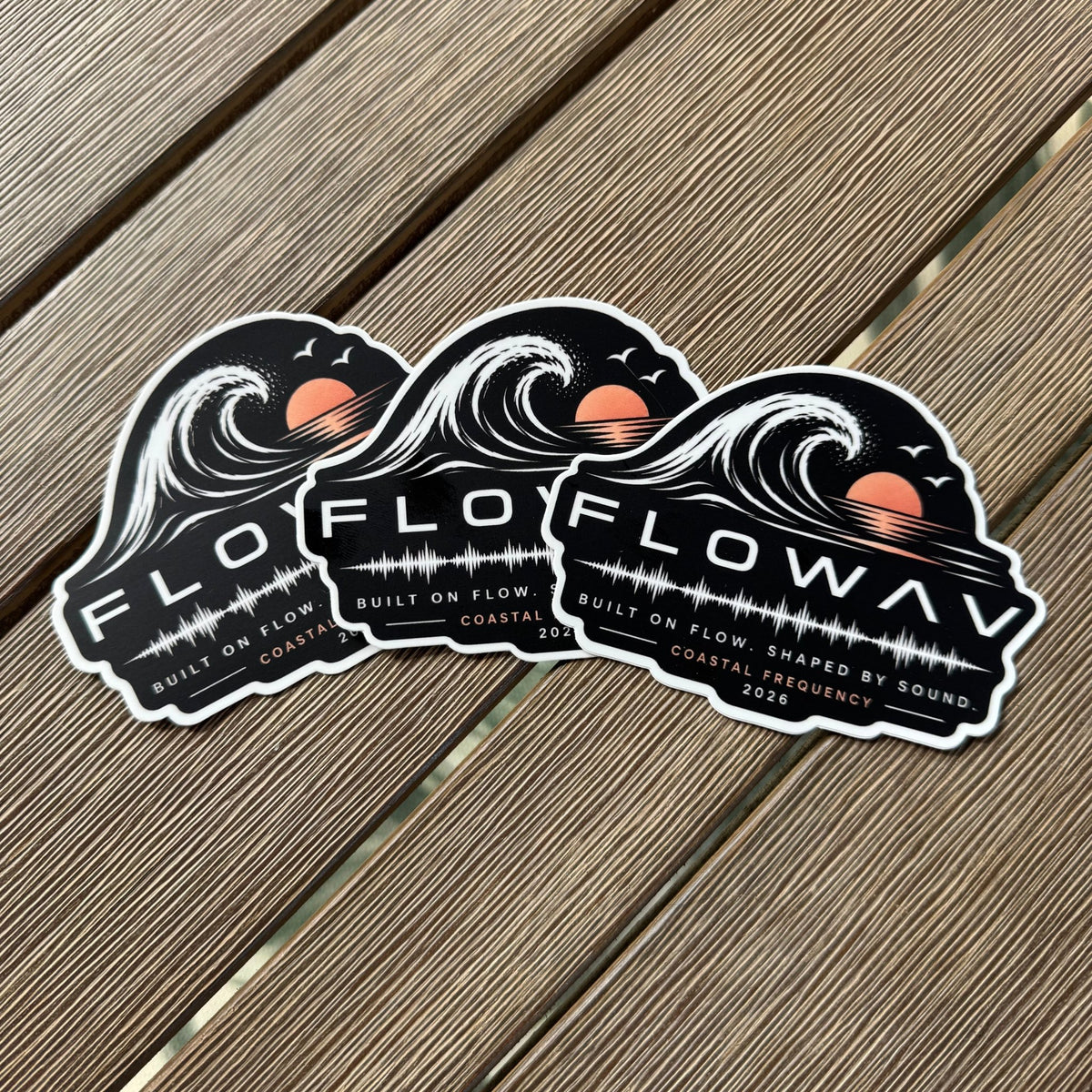 Three identical Flowav Coastal Frequency die-cut stickers featuring a stylized ocean wave, sunset, and soundwave graphic, displayed on a brown wooden table.