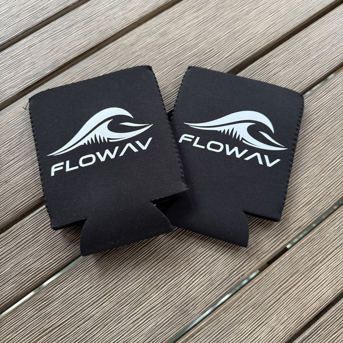 Two black FLOWAV neoprene can coolers with white wave logos on an outdoor wood table.