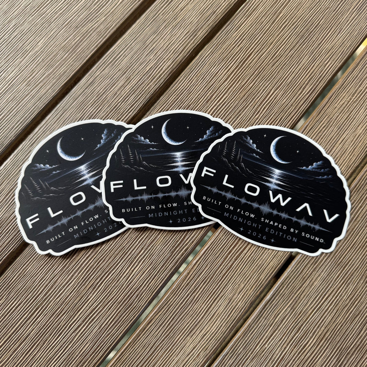 Three identical FLOWAV Midnight Edition vinyl stickers are arranged in a fan shape on a brown wooden plank surface. The stickers feature a dark night scene with a crescent moon, stars, and a soundwave, with the text 'FLOWAV MIDNIGHT EDITION' and 'BUILT ON FLOW, SHAPED BY SOUND'.