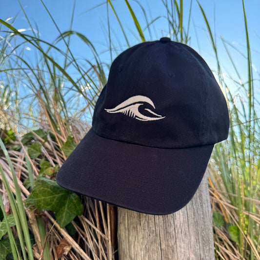 FLOWAV Richardson R55 classic black baseball hat with cream embroidered wave logo resting on a weathered beach fence post among coastal dunes