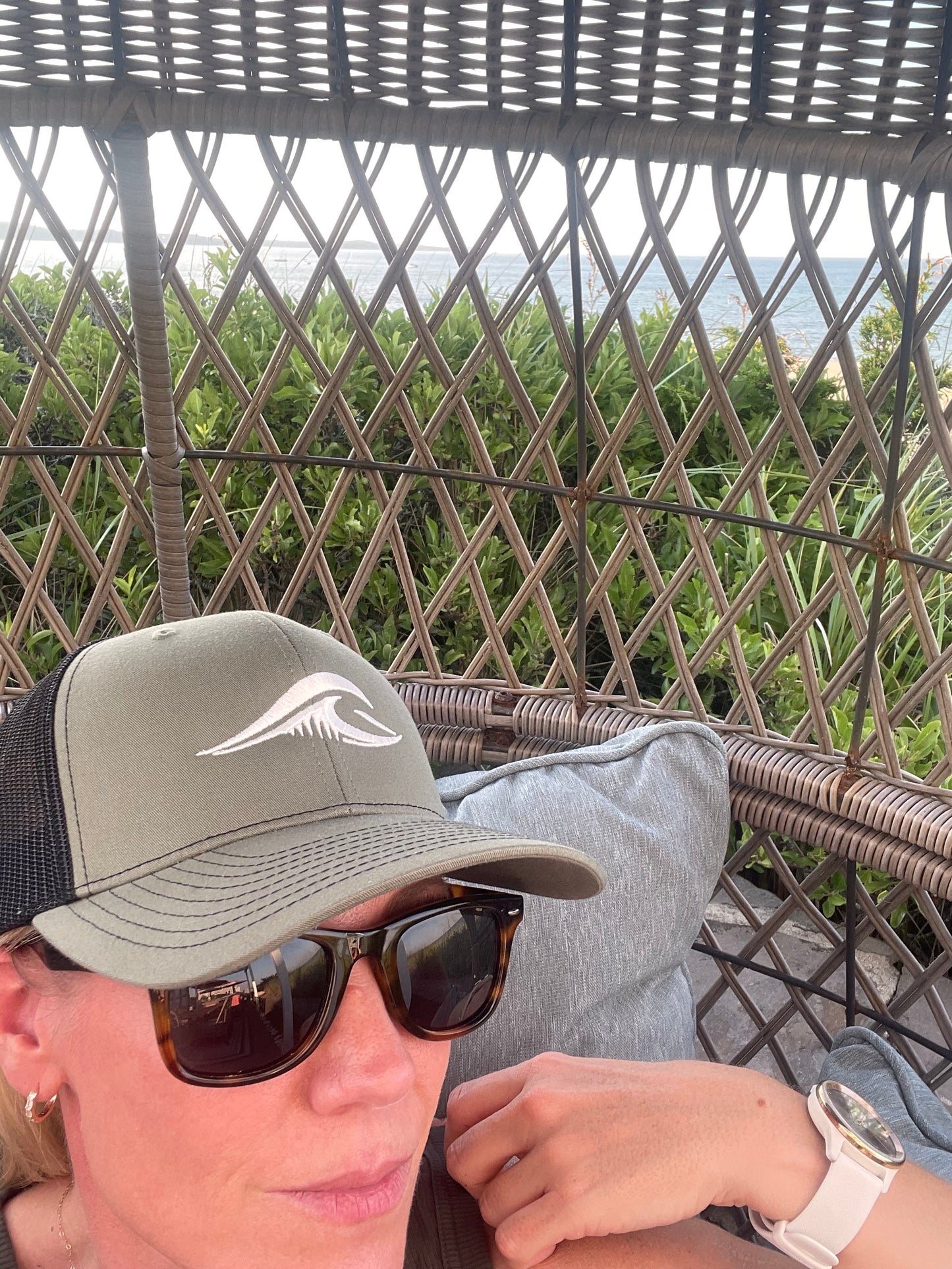 Person wearing a loden black FLOWAV Richardson 112 Trucker Hat with cream embroidered logo while relaxing in a woven beach cabana.