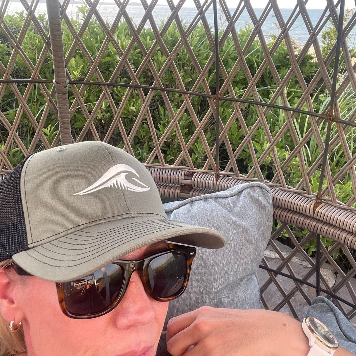 Person wearing a loden black FLOWAV Richardson 112 Trucker Hat with cream embroidered logo while relaxing in a woven beach cabana.