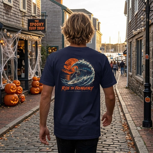 A man wearing the FLOWAV Ride the Freakuency Graphic Tee in True Navy in a coastal Halloween town showing the skeleton surfer