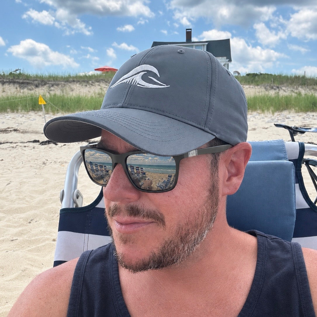 A man wearing a charcoal gray baseball cap with a white FLOWAV wave logo, black sunglasses, and a black tank top sitting in a beach chair at the sand dunes under a blue sky.