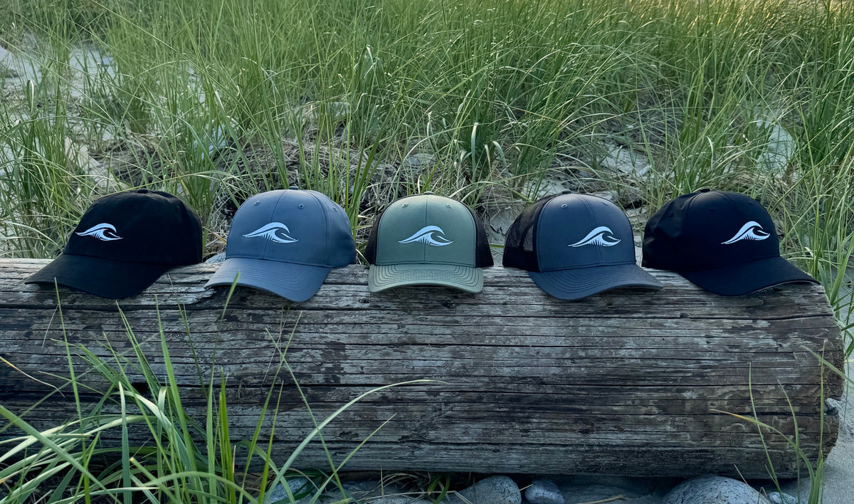 five flowav hats on weathered beachwood including black R55, charcoal Richardson R225, loden Richardson 112 classic trucker, charcoal 112 trucker and black Richardson R225 with seagrass in the background.
