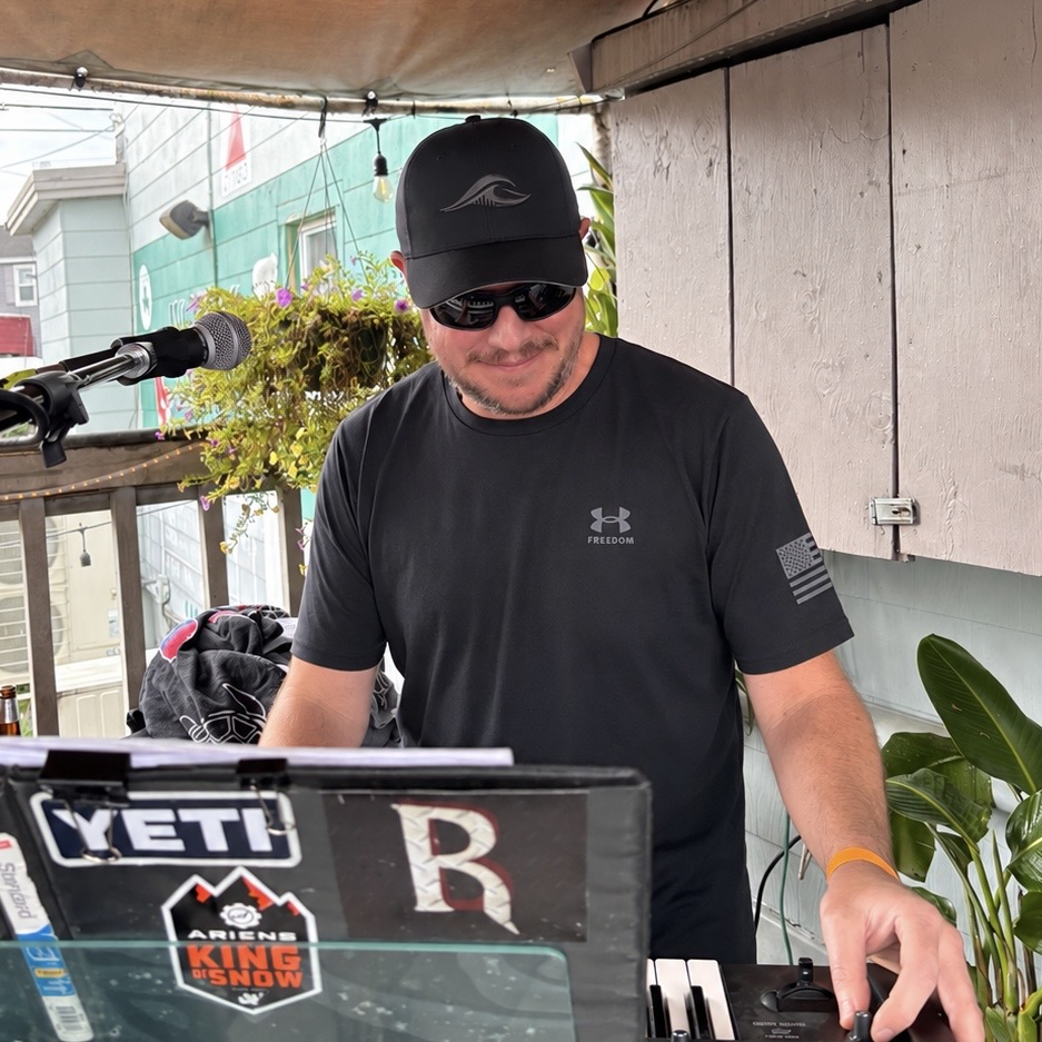 A man wearing a black Under Armour t-shirt and a black FLOWAV Midnight Edition Richardson R225 hat stands behind a keyboard and a microphone at a live music outdoor event, with a YETI sticker in the foreground and a patio canopy overhead.