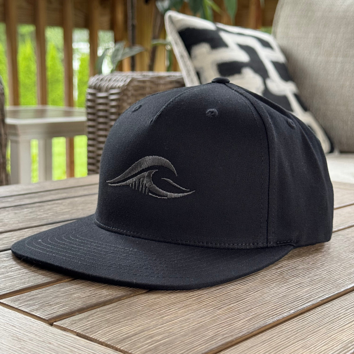 A black flat-brim baseball cap with a dark gray embroidered wave logo, resting on a wooden table in an outdoor patio setting. FLOWAV Midnight Edition.