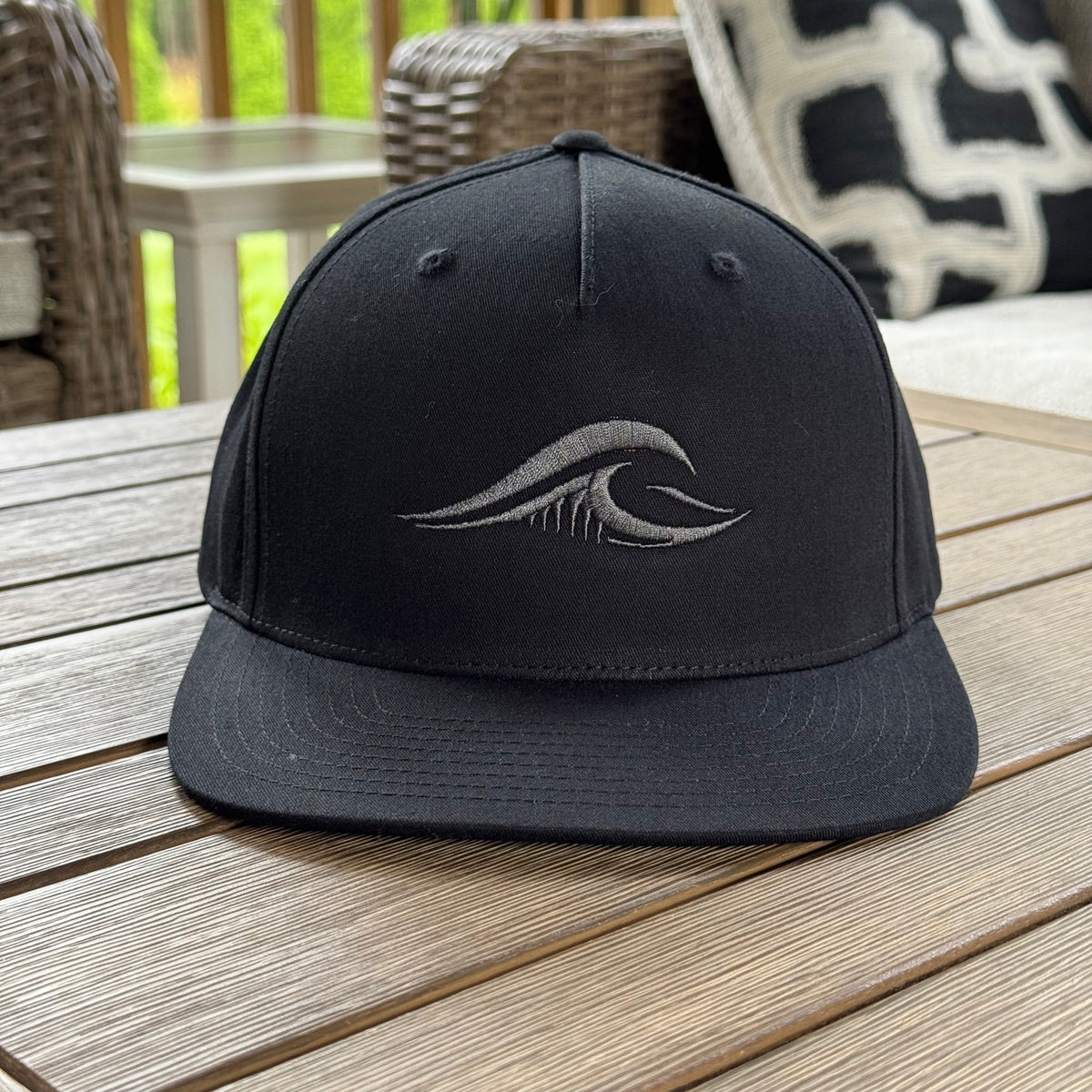A black FLOWAV Midnight Edition Richardson R255 flat-brim baseball cap with a dark gray tonal embroidered wave logo on the front, resting on a wooden deck table with outdoor patio furniture in the background.
