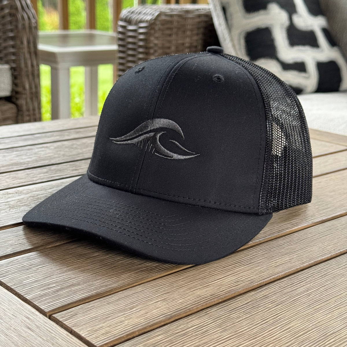 A black trucker-style baseball cap with a curved brim and black mesh back panels, featuring a FLOWAV embroidered dark gray wave logo on the front, displayed on a wooden outdoor table.