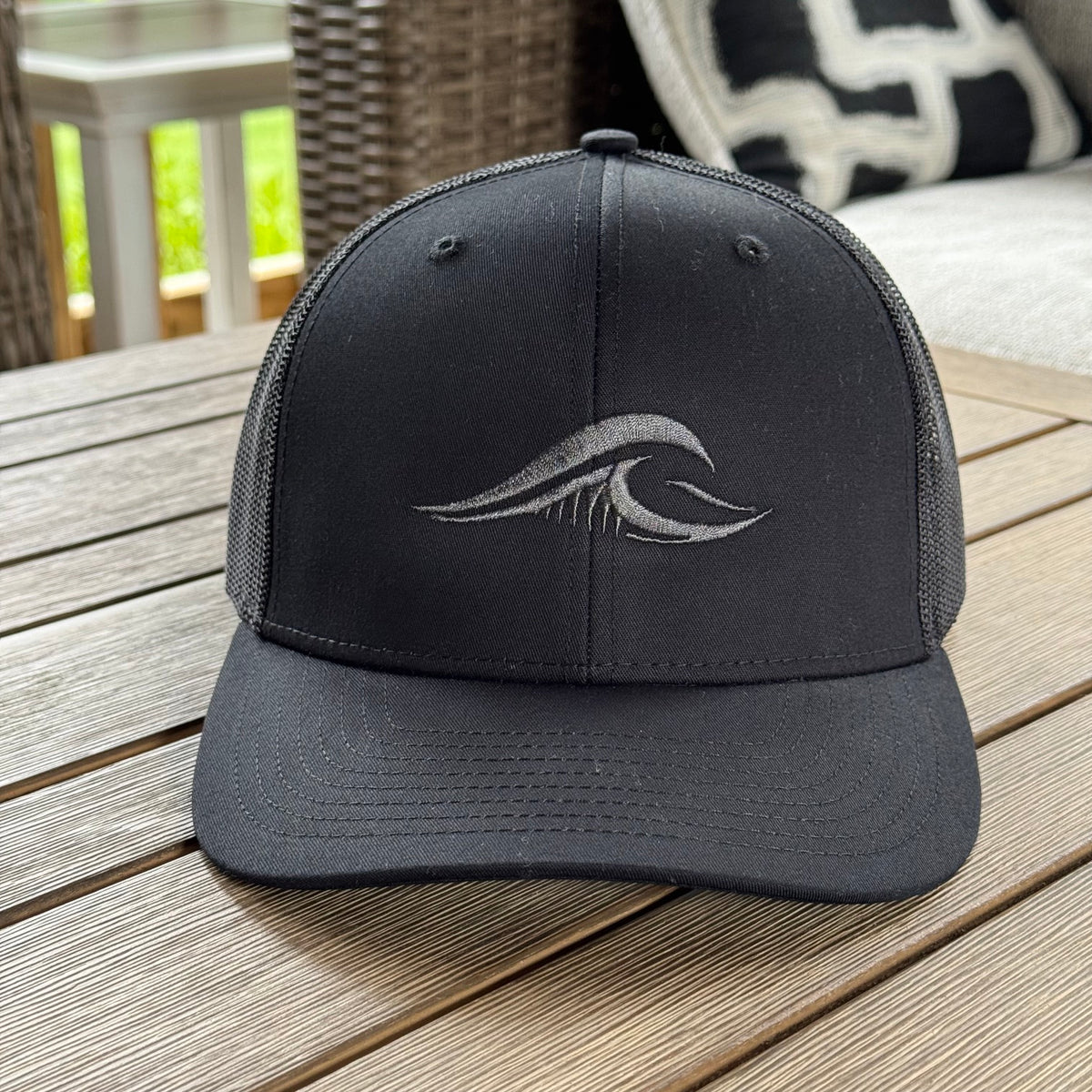 A FLOWAV black Richardson 112 classic trucker hat with a tonal black logo on the front sitting on a weathered wood table.