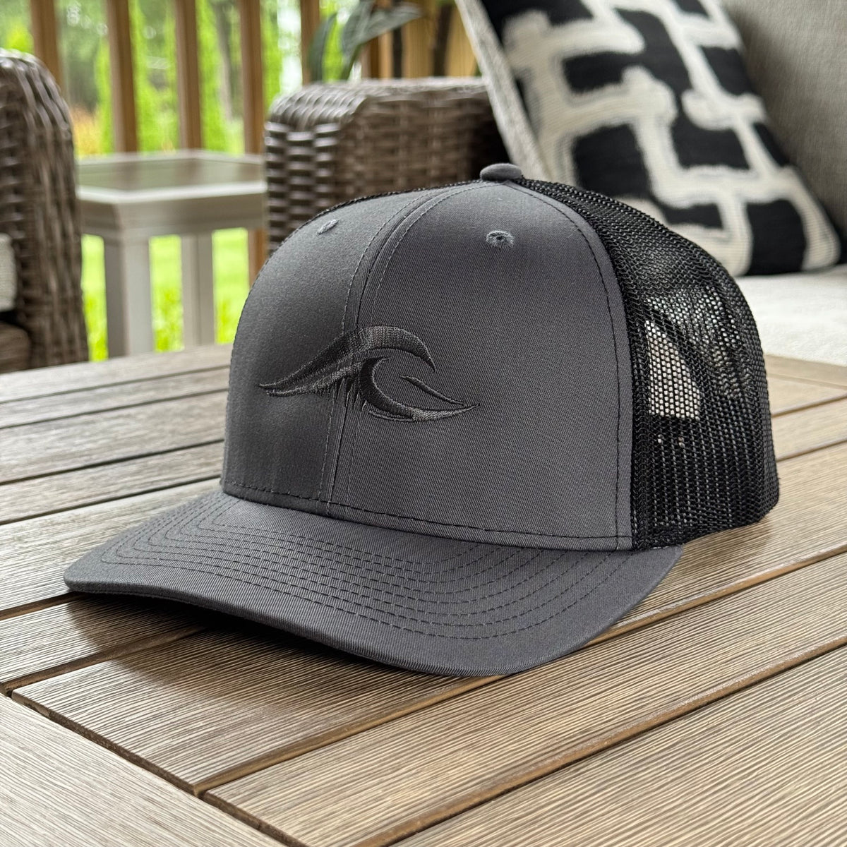 A charcoal gray FLOWAV Richardson 112 Trucker Hat featuring a tonal gray wave logo angled on a weathered wood outdoor table.