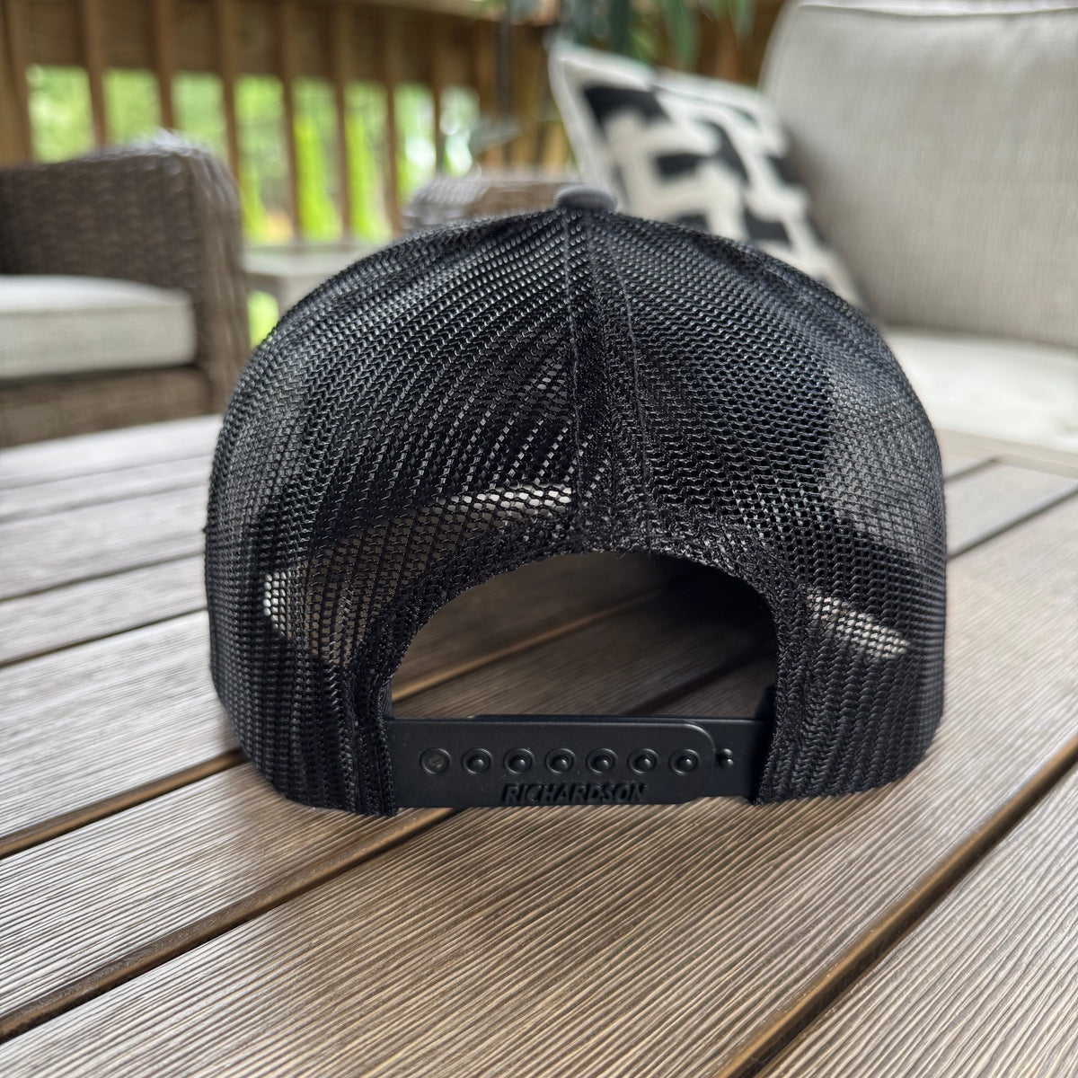 A black mesh-back trucker cap viewed from the back showing the black mesh and richardson 112 snapback, resting on a wooden deck table with patio furniture in the background.