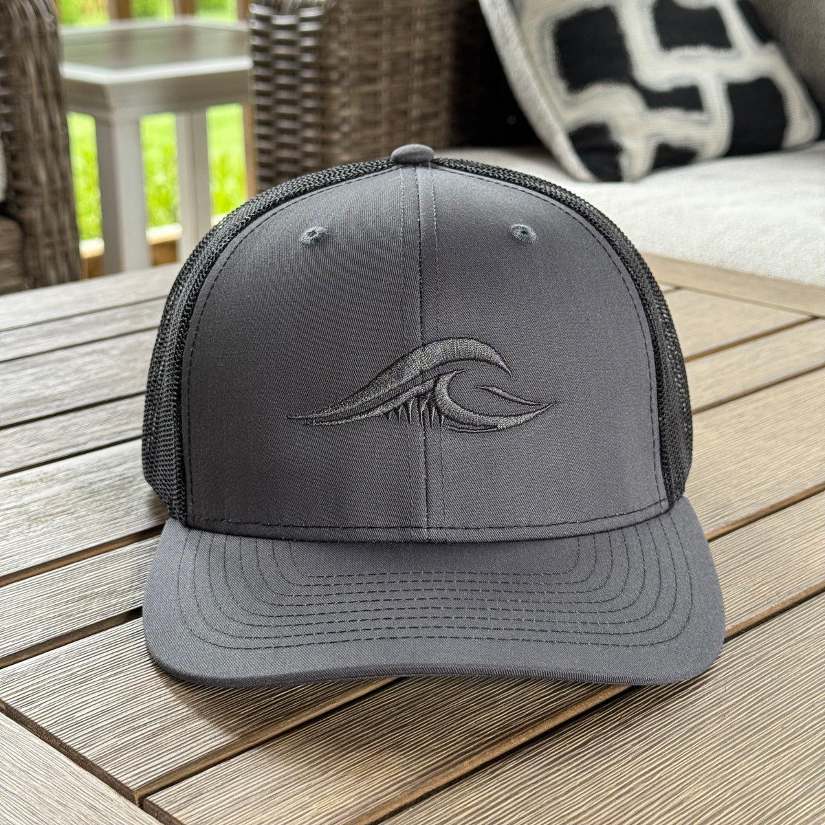 A dark gray FLOWAV Richardson 112 Trucker hat featuring charcoal tonal wave logo on the front resting on a wooden outdoor table.