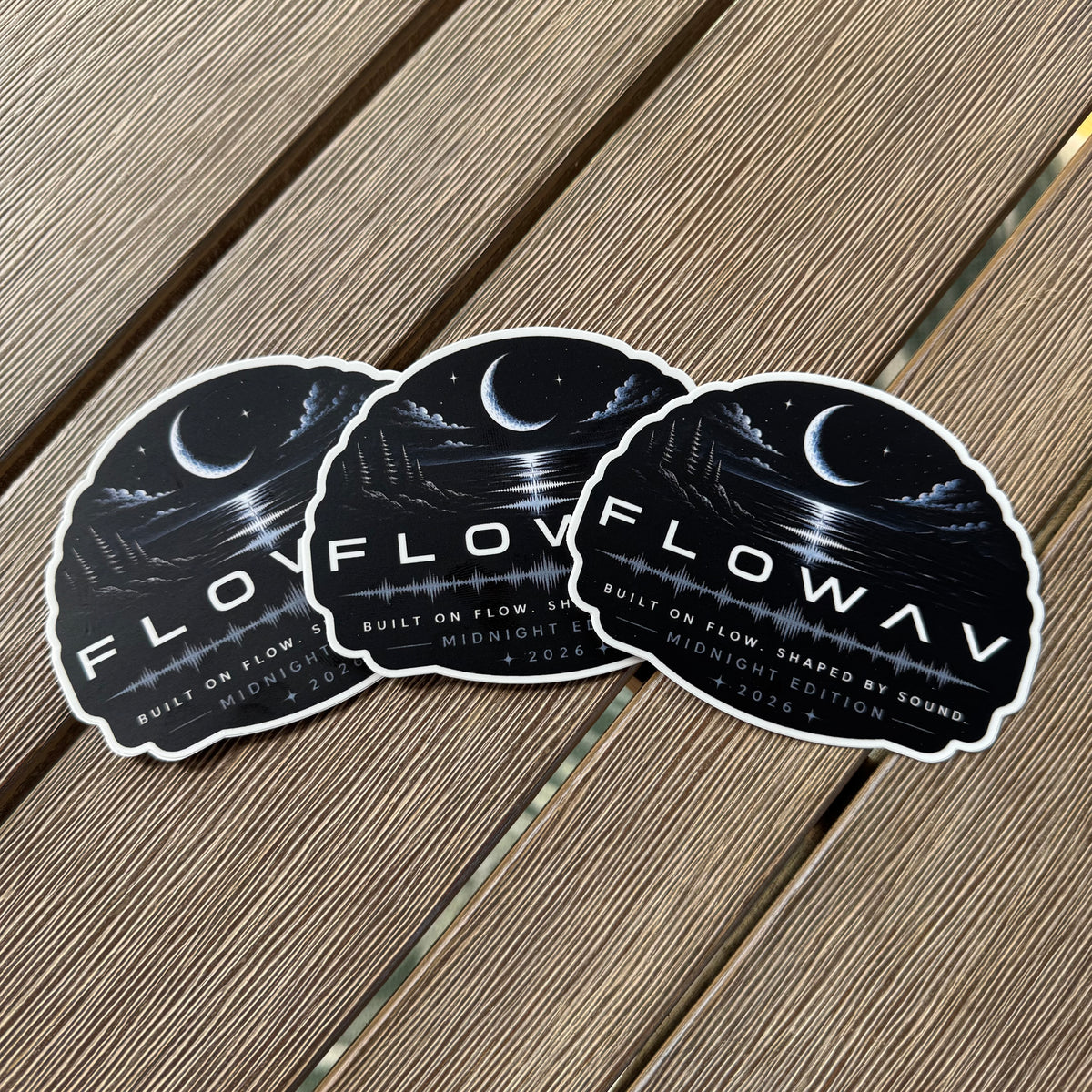 Three identical FLOWAV Midnight Edition vinyl stickers are arranged in a fan shape on a brown wooden plank surface. The stickers feature a dark night scene with a crescent moon, stars, and a soundwave, with the text 'FLOWAV MIDNIGHT EDITION' and 'BUILT ON FLOW, SHAPED BY SOUND'.