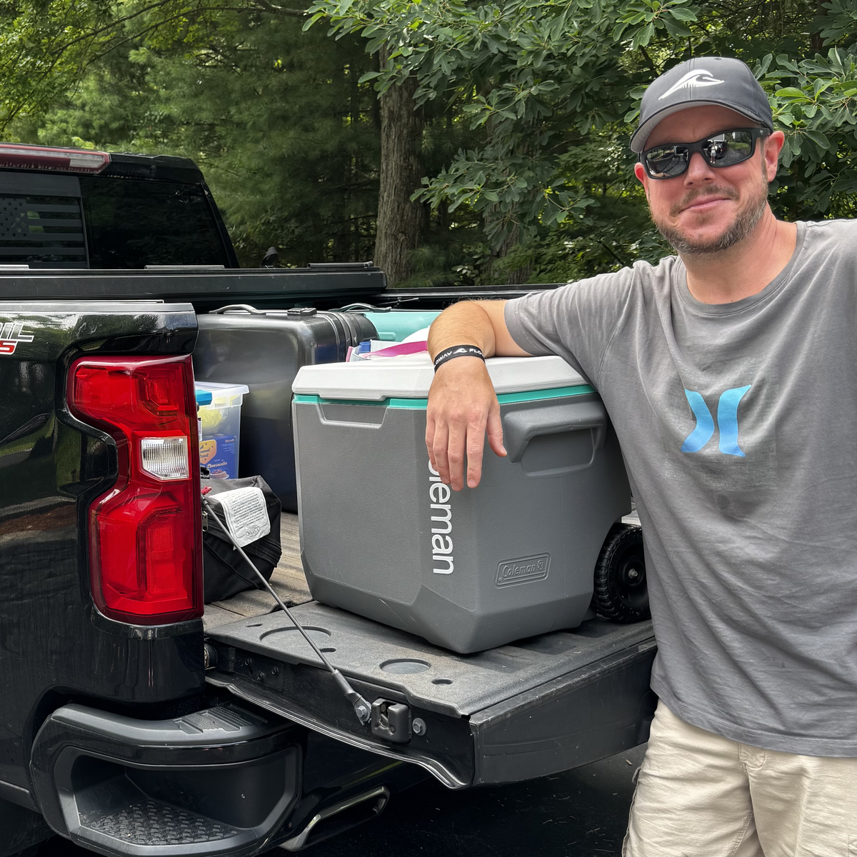 A man wearing a gray FLOWAV Richardson R225 hat and a gray Hurley t-shirt and sunglasses stands next to a black pickup truck tailgate, holding a gray Coleman cooler.