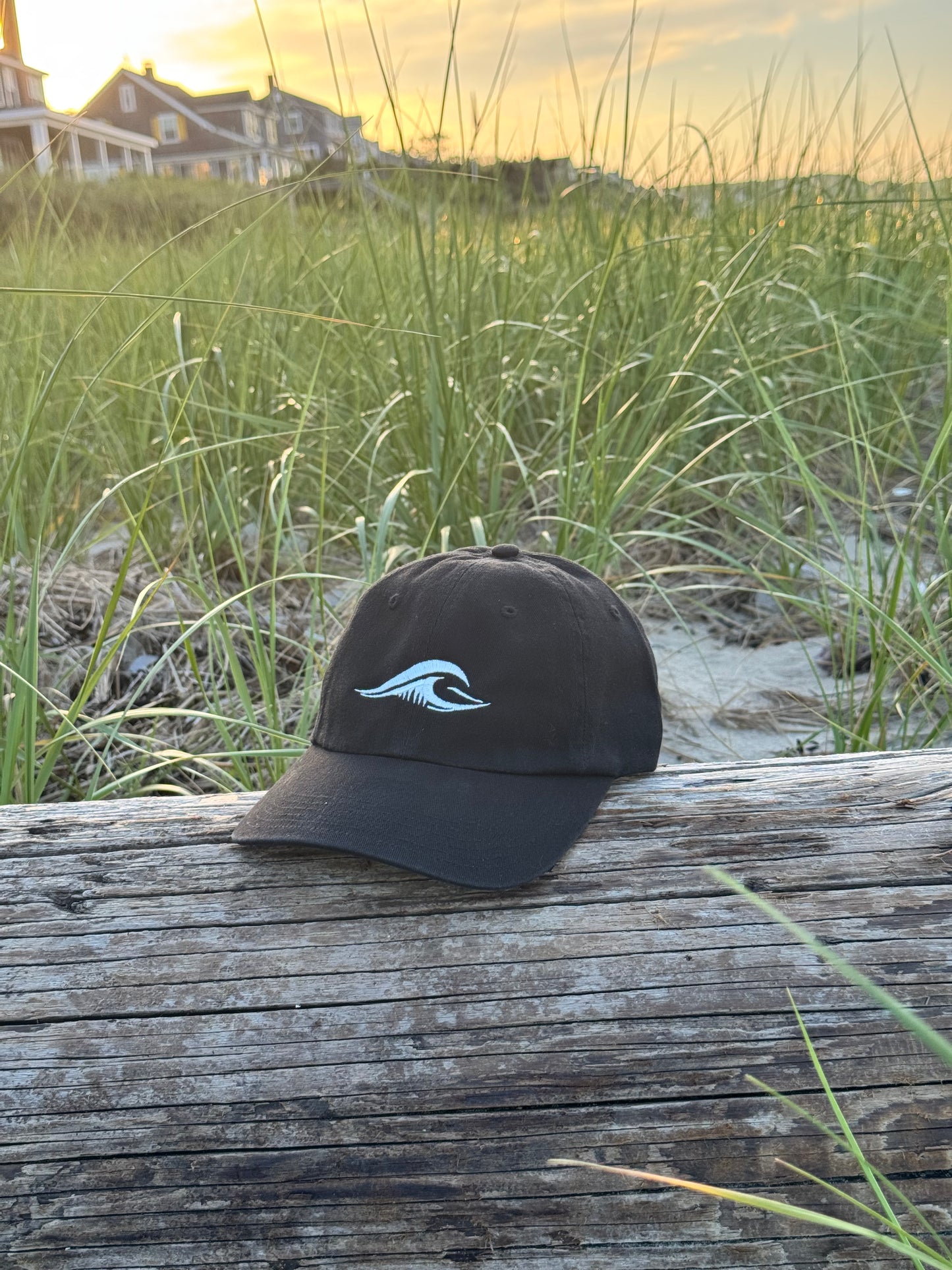 FLOWAV Richardson R55 black baseball hat with cream embroidered wave logo resting on weathered driftwood at the beach during sunset.