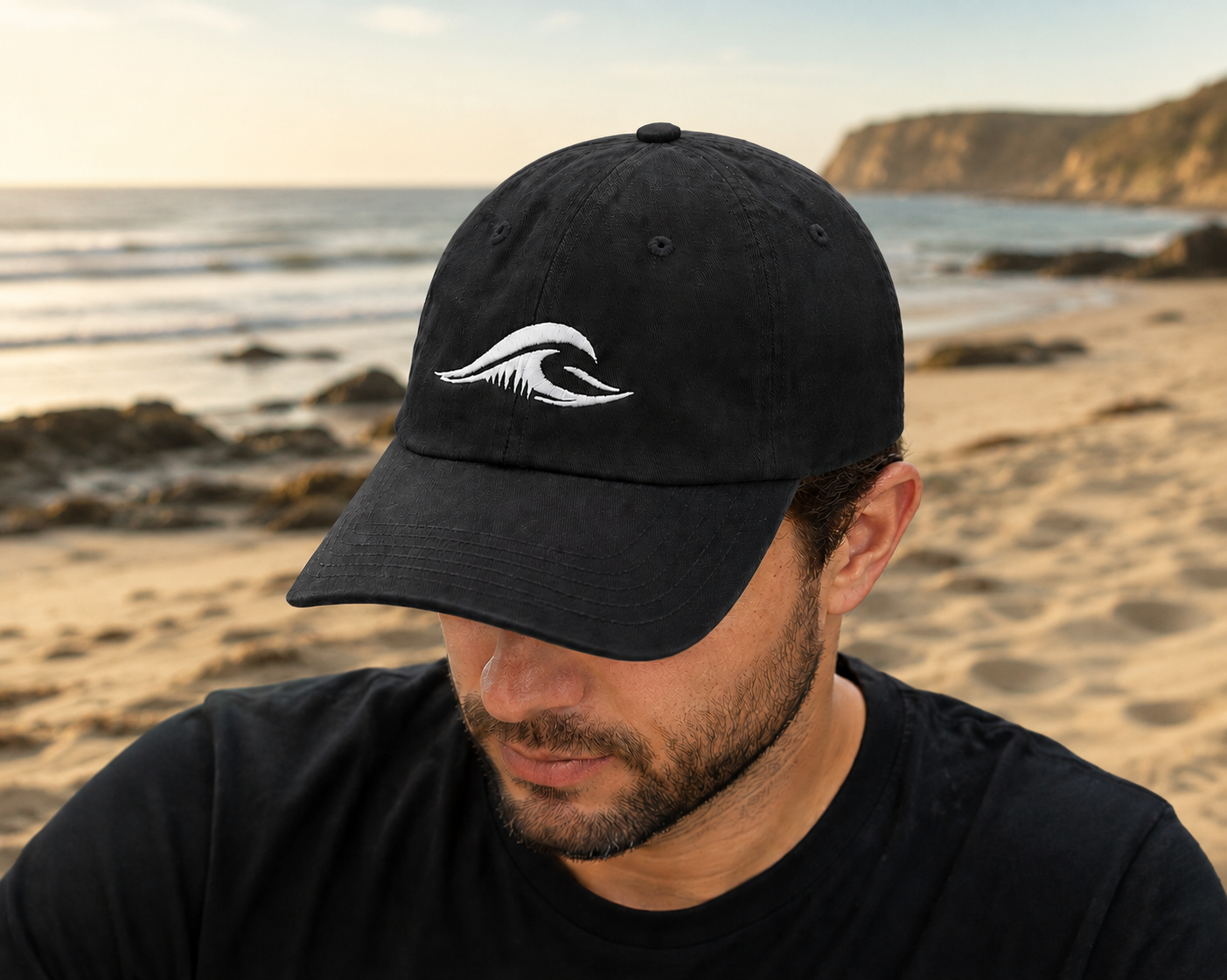 Person wearing a FLOWAV Richardson R55 classic black baseball hat with cream embroidered wave logo on a beach at sunset