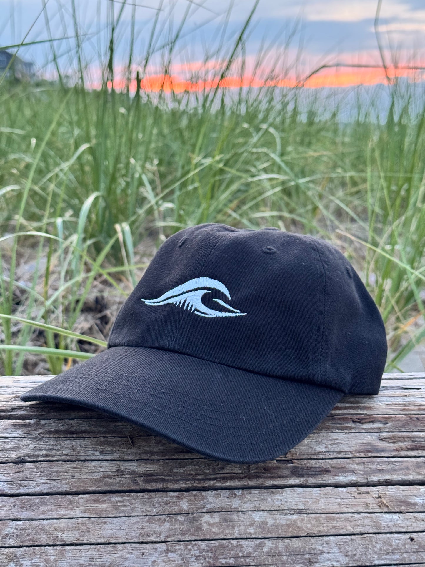 FLOWAV Richardson R55 classic black baseball hat with cream embroidered wave logo resting on weathered beach wood with coastal dunes and sunset in the background.