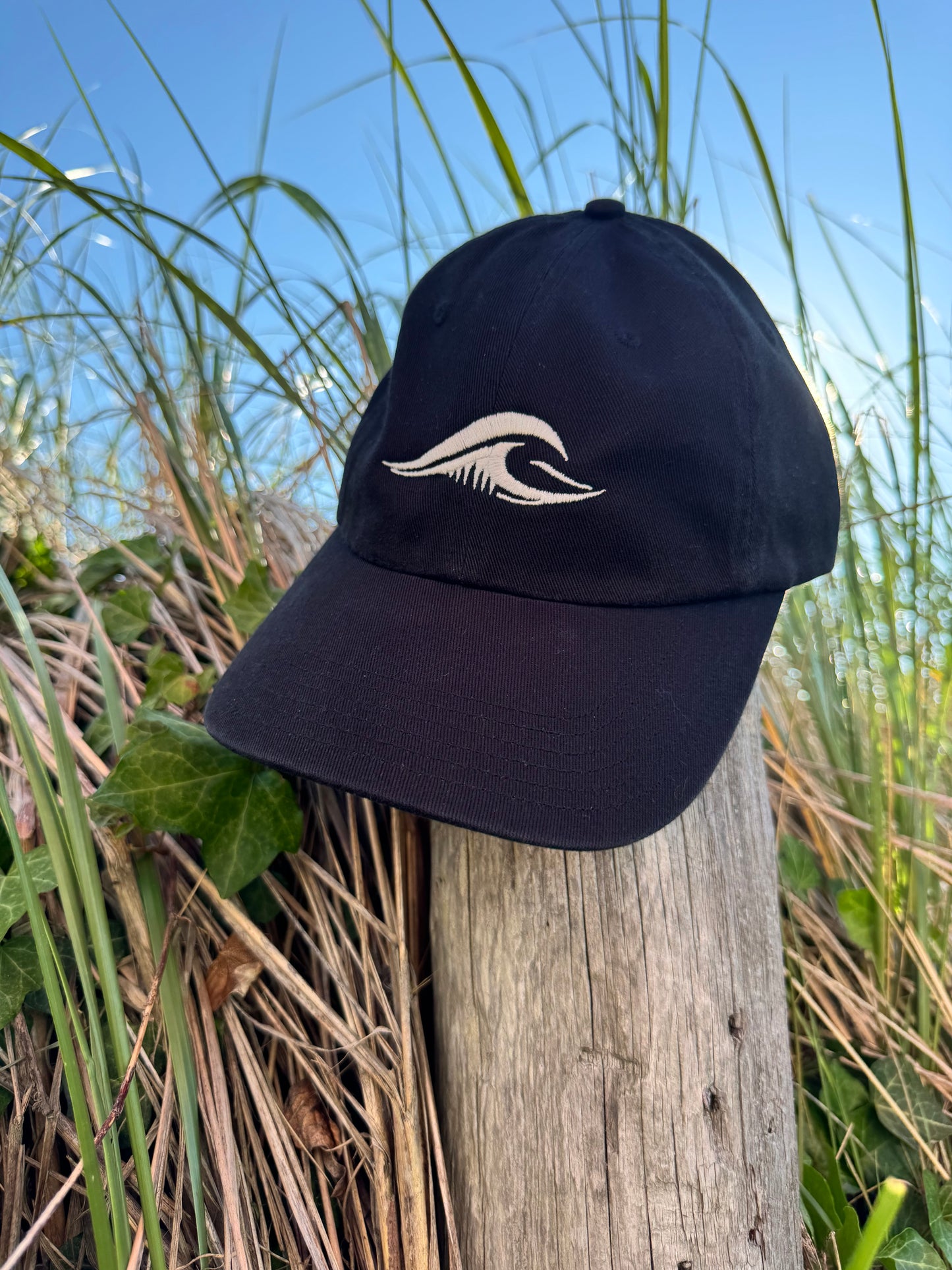 FLOWAV Richardson R55 classic black baseball hat with cream embroidered wave logo resting on a weathered beach fence post among coastal dunes