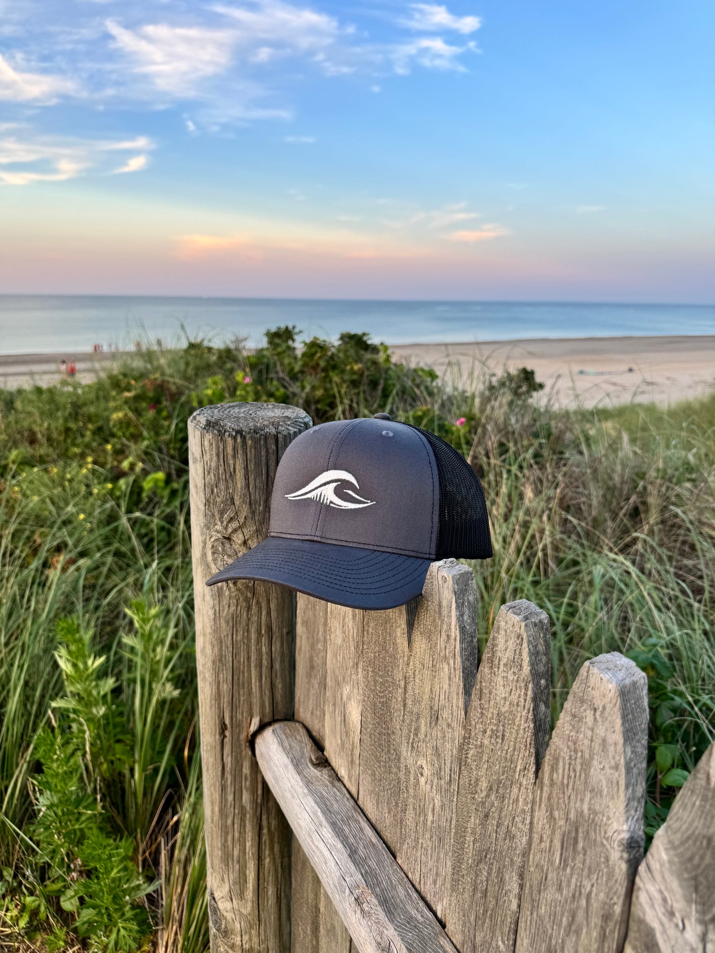 Charcoal FLOWAV Richardson 112 trucker hat on a weathered beach fence with an ocean sunset in the background.