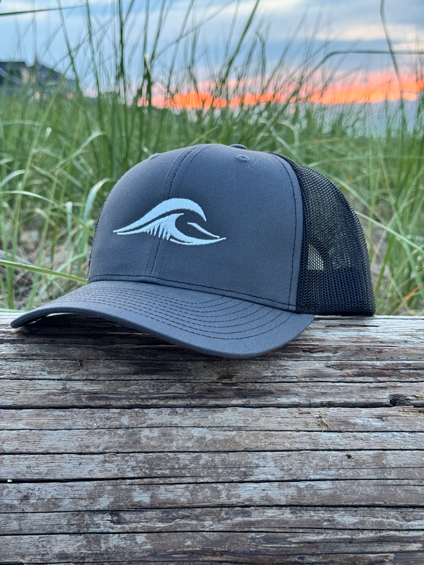 FLOWAV Richardson 112 Charcoal Trucker Hat with cream embroidered logo resting on weathered driftwood overlooking the beach at sunset