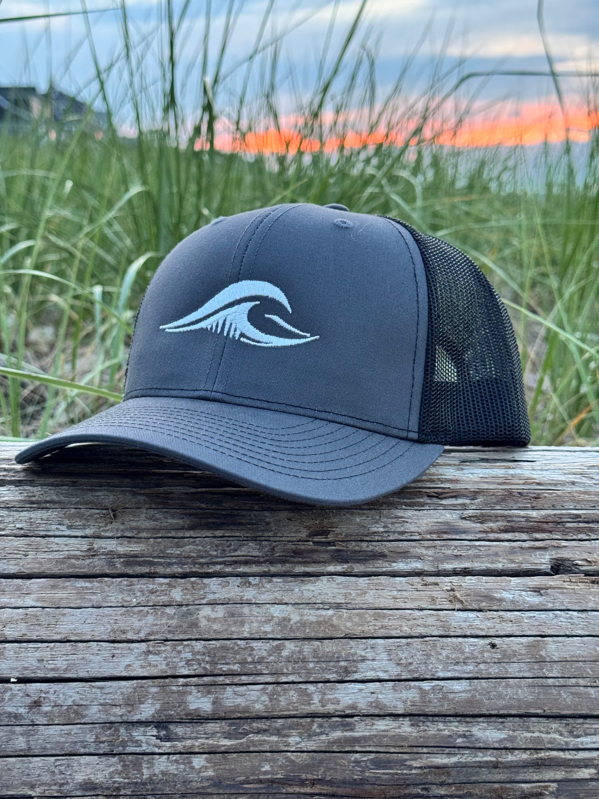 FLOWAV Richardson 112 Charcoal Trucker Hat with cream embroidered logo resting on weathered driftwood overlooking the beach at sunset
