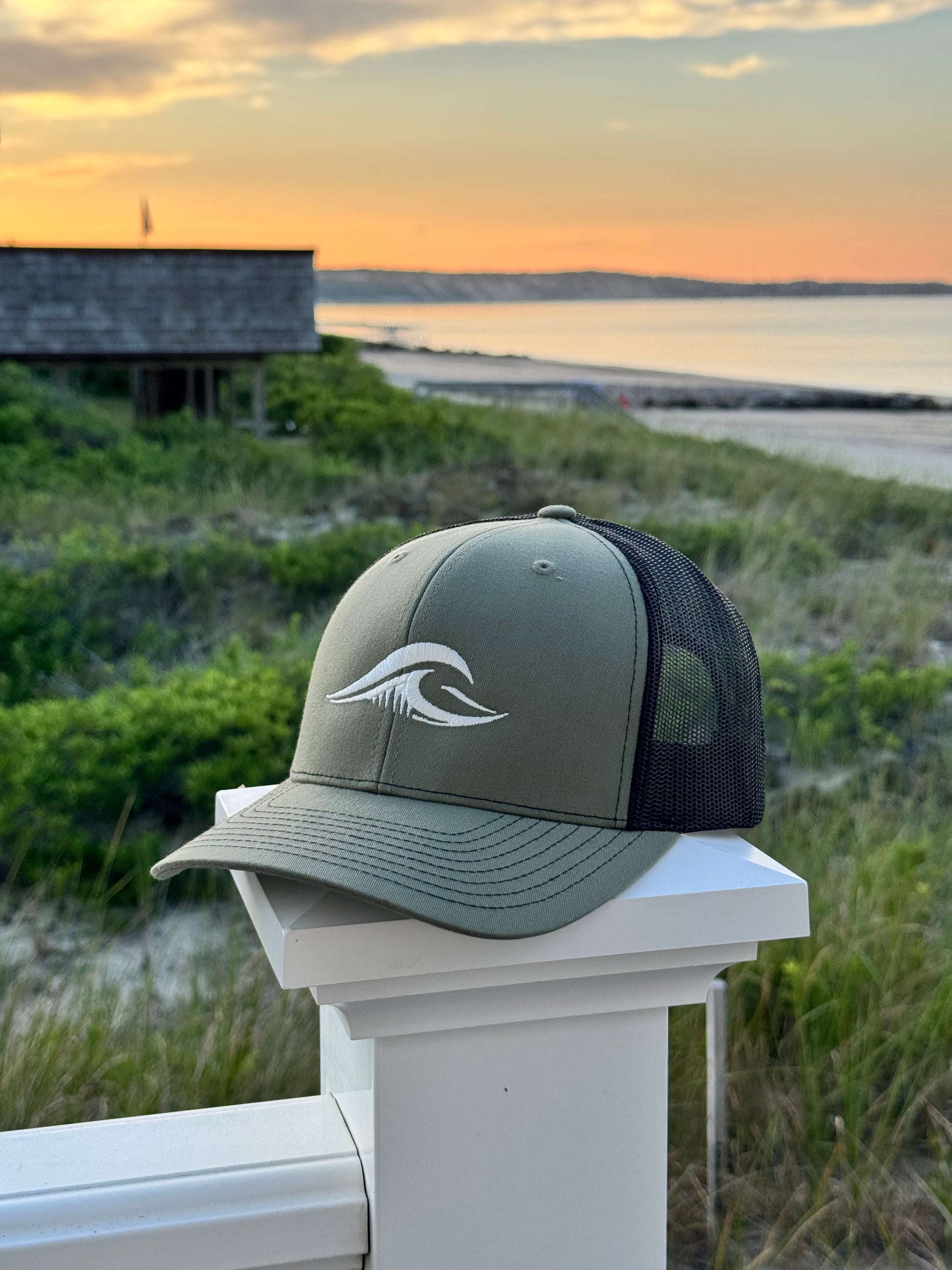 Loden Black FLOWAV Richardson 112 trucker hat with cream embroidered wave logo on a white beach fence overlooking the ocean at sunset