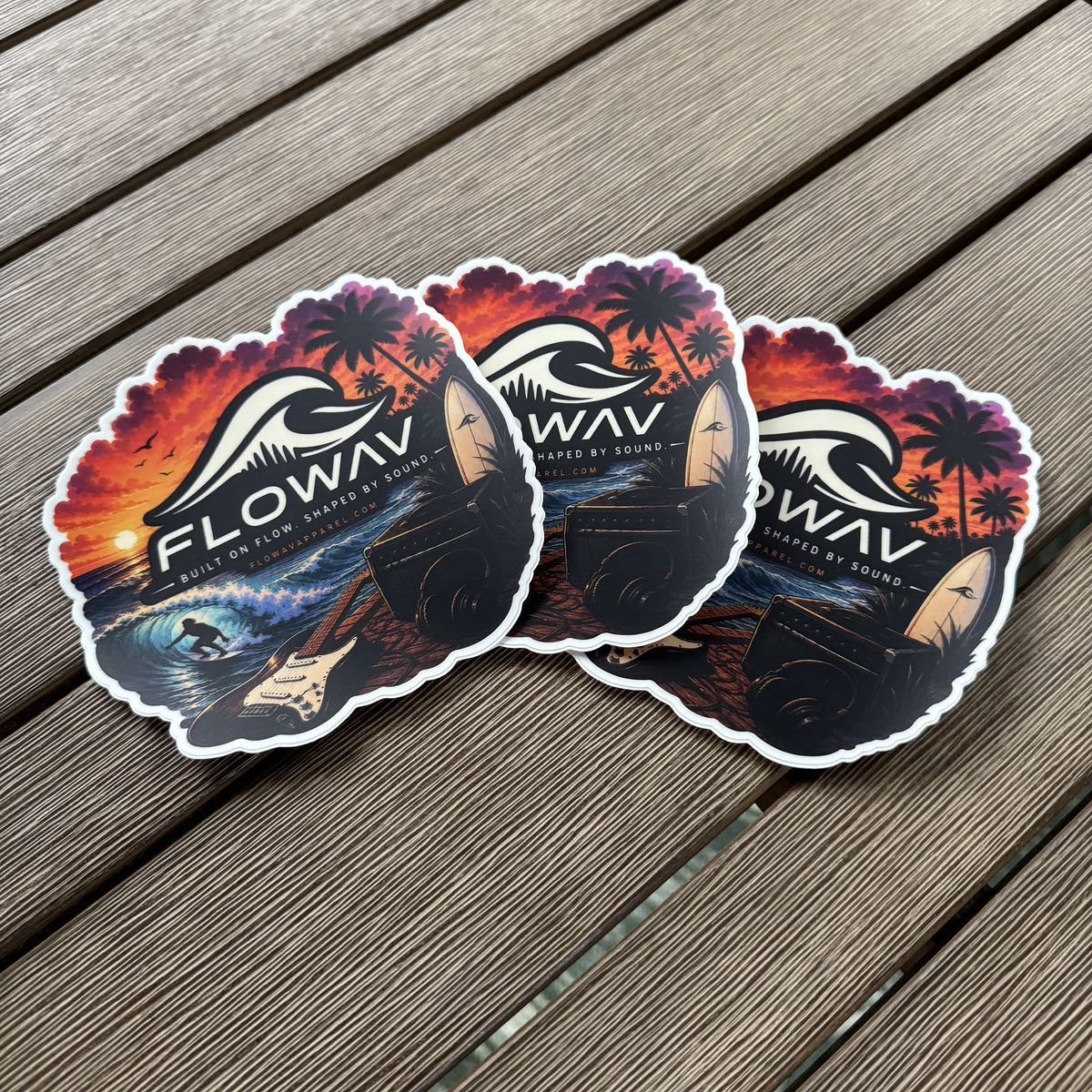 Three die-cut FLOWAV Sunset Stickers featuring a guitar, amp, surfer on a breaking wave, sunset, surfboard and headphones on a weathered wood deck.