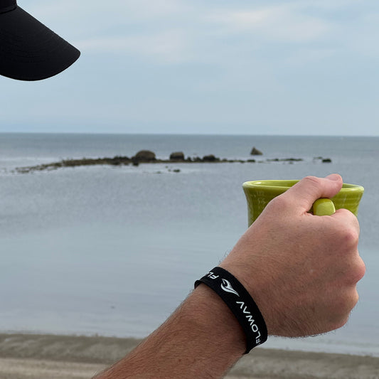 A person outdoors by the ocean water holding a lime green mug, wearing a black cloth wristband with white FLOWAV text and wave logo. The background shows a calm sea and cloudy sky.
